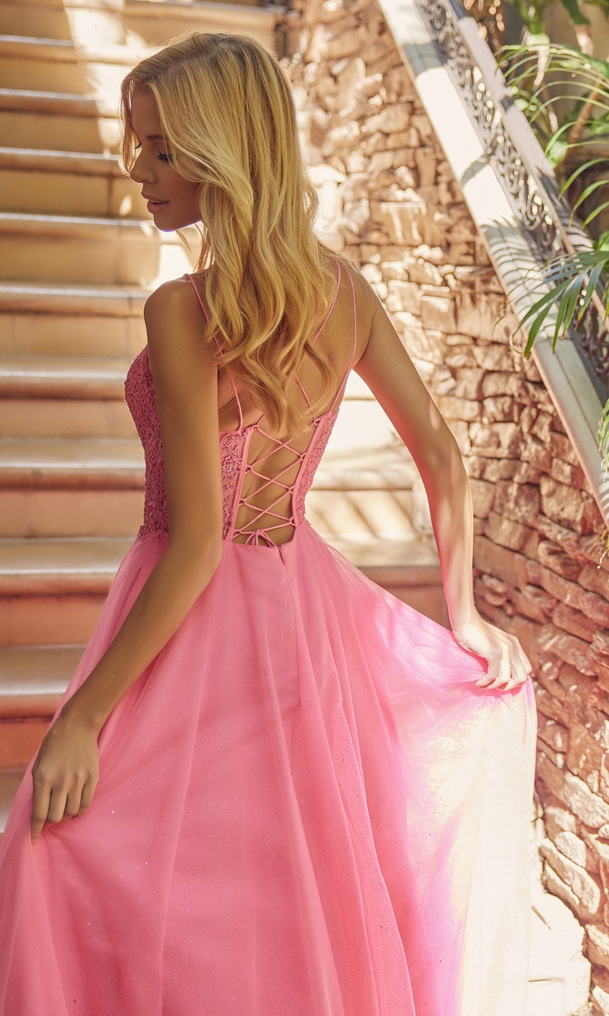 A woman in the Glitter-Tulle Long Corset-Back Prom Ball Gown 268 stands on a staircase, looking over her shoulder and holding part of the dress.